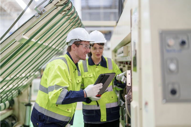 A field inspector and a colleague wearing hard hats and high-visibility safety vests using a mobile tablet to fill out a digital safety inspection checklist in a manufacturing facility.