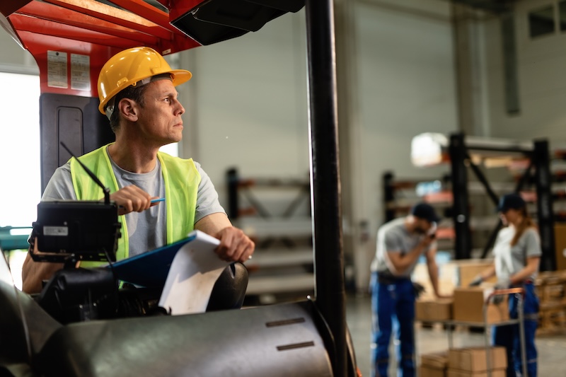 Warehouse forklift operator reviewing a paper clipboard, highlighting the need for digital inspection forms in manufacturing and transport logistics